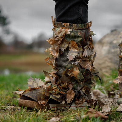 A person wearing brown leaf suit gaiters attached to boots, blending into a leafy camouflage environment.