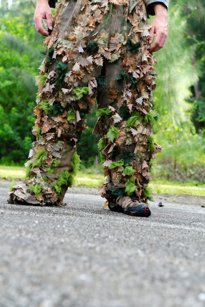 A person wearing camouflage leafy ghillie pants and hiking shoes standing on a paved path outdoors with green foliage in the background.
